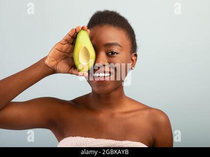 Bonne jeune femme afro-américaine tenant des moitiés fraîches d'avocat et couvrant l'œil, souriant à l'appareil photo sur fond gris Banque D'Images