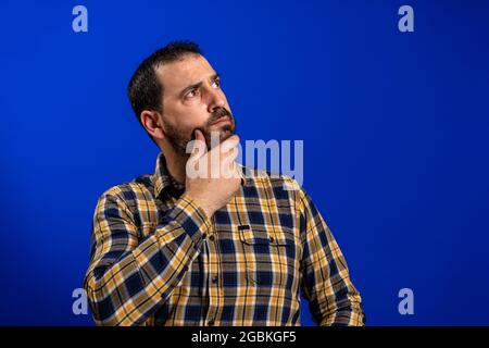 Frognant homme pensant exprimant des doutes et des préoccupations. Petit garçon avec barbe en chemise à carreaux bleu isolée sur fond bleu studio Banque D'Images