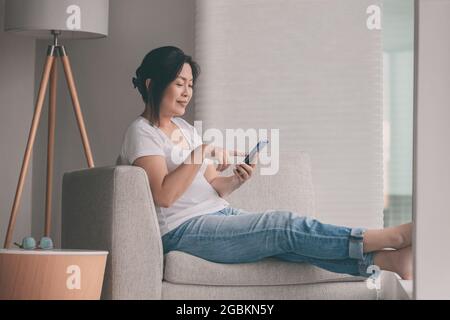 Travail à domicile une femme asiatique mûre heureuse se détendant dans un canapé en utilisant le téléphone mobile shopping en ligne ou le travail à distance Banque D'Images