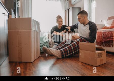 Journée de fête mouvante. Couple de Yong se déplacer dans la nouvelle maison. Jeune couple heureux, jeunes mariés qui boivent du vin en famille et qui s'amusent dans la chambre avec des boîtes en carton Banque D'Images