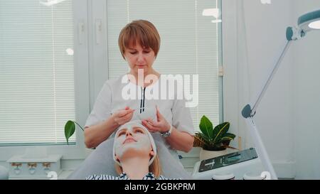 Esthéticienne mettant le masque crème sur le visage de la femme au salon de beauté. Soins du visage. Banque D'Images