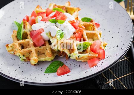 Gaufres de courgettes, beignets de courgettes de courgettes cuisant sur un gaufrier, gaufres de courgettes végétariennes avec tomates, crème aigre et basilic dans une assiette blanche. Banque D'Images