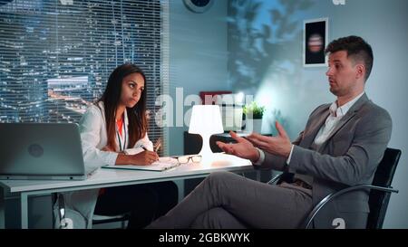 Session de thérapie psychologue: Homme d'affaires partageant ses problèmes avec un spécialiste de la santé psychologique dans un bureau moderne confortable en soirée Banque D'Images