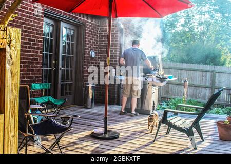 Homme avec un verre de vin de cuisine au barbecue extérieur avec beaucoup de fumée s'affichant sur le patio avec des chaises et chat et parapluie rouge Banque D'Images