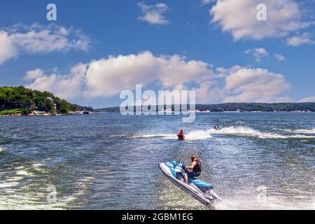 Les gens sur les motomarines sautent le réveil du bateau sur le lac avec des maisons sur la rive au loin. Banque D'Images