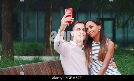 Jeune couple joyeux prenant selfie assis sur un banc à l'extérieur. Ils passent leur temps dans le grand parc. Banque D'Images