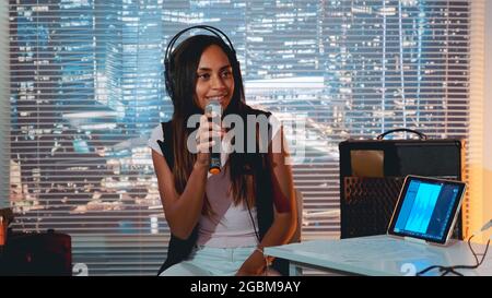 Jolie fille noire dans casque chantant dans le microphone et enregistrer une chanson dans le studio à la maison avec gratte-ciel en arrière-plan. Banque D'Images