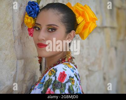 Jeune magnifique danseuse folklorique mexicaine Yucatecan porte une robe folklorique traditionnelle avec des fleurs colorées dans ses cheveux et regarde par-dessus son épaule. Banque D'Images