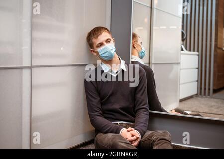 Un type de nationalité caucasienne est assis dans un coin portant un masque protecteur triste au miroir. Banque D'Images