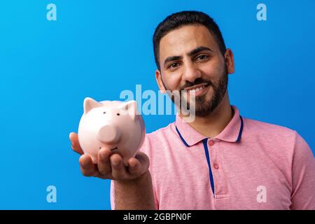 Beau homme hispanique très excité tenant la banque de porc contre fond bleu Banque D'Images