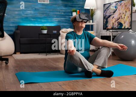 Femme sénior en bonne santé utilisant un casque de réalité virtuelle méditant tout en étant assise en position lotus sur le tapis de yoga dans le salon pendant l'entraînement de méditation. Pensioner entraînement corps muscle faire des sports exercice Banque D'Images