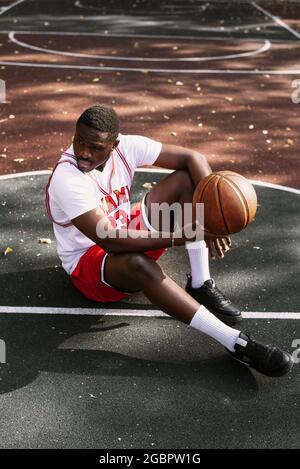 Portrait d'un jeune afro-américain tenant un ballon de basket et assis sur un terrain de basket-ball. Faites une pause pendant votre entraînement. Sport tendance Banque D'Images