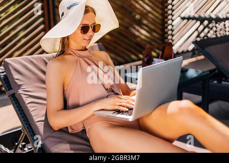 Nous travaillons même en vacances, belle jeune femme en lunettes de soleil et un chapeau, se trouve sur un transat près de la piscine avec un ordinateur portable, la femme utilise un ordinateur portable sur va Banque D'Images