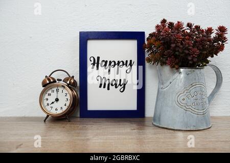 Texte typographique Happy May avec réveil et décoration artificielle de plantes sur table en bois et fond de mur blanc Banque D'Images