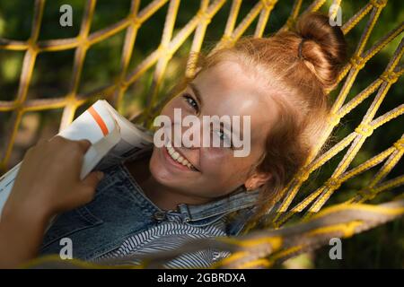 Jeune femme blonde étudiante souriante lisant UN livre tout en étant allongé sur UN hamac dans le jardin. Femme se détendant dans un hamac. Retour à l'école, campus universitaire Banque D'Images