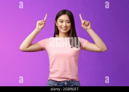 Adorable femme asiatique aux cheveux sombres et affirmée porter un t-shirt rose lever les mains, montrer la promotion, pointer les doigts vers le haut, sourire, inviter utiliser le lien, essayez Banque D'Images