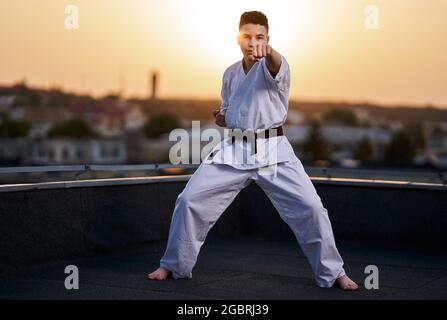 Jeune adolescent karaté praticien en kimono blanc entraînement sur le toit au coucher du soleil Banque D'Images