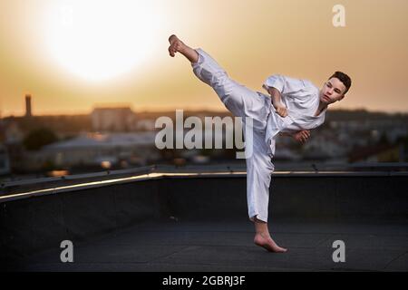 Jeune adolescent karaté praticien en kimono blanc entraînement sur le toit au coucher du soleil Banque D'Images