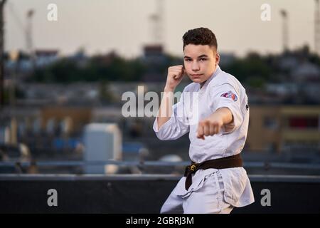 Jeune adolescent karaté praticien en kimono blanc entraînement sur le toit au coucher du soleil Banque D'Images