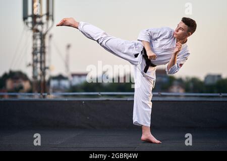 Jeune adolescent karaté praticien en kimono blanc entraînement sur le toit au coucher du soleil Banque D'Images