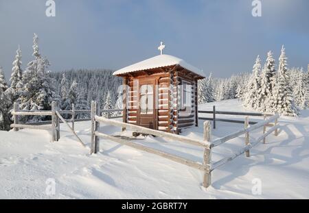 Froid jour d'hiver. Sur la pelouse il y a une église en bois couverte de neige sur les montagnes et autour il y a une forêt avec de beaux arbres. Banque D'Images