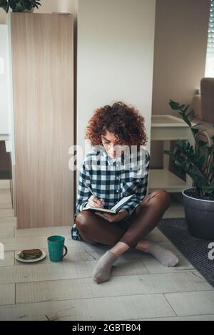 Charmante femme aux cheveux bouclés écrit quelque chose dans un livre tout en travaillant et en buvant un café avec un sandwich Banque D'Images