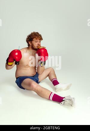 Drôle d'homme à tête rouge dans des shorts de boxe bleus et des gants isolés sur fond gris de studio. Concept de sport, d'humour et de corps positif. Banque D'Images