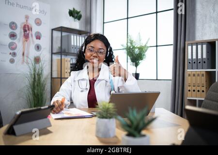 Femme afro-américaine souriante médecin généraliste en manteau blanc et écouteurs casque, assis sur le lieu de travail dans la clinique, en utilisant un ordinateur portable pour écrire la prescription de patient ou de prendre des notes, montrant le pouce vers le haut Banque D'Images