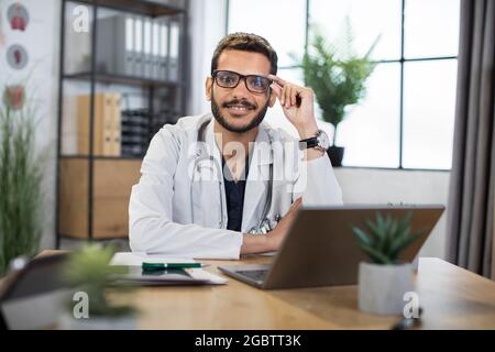 Gros plan d'un beau médecin malais de 30 ans souriant, assis à la table avec un ordinateur portable dans son bureau privé et regardant l'appareil photo. Banque D'Images