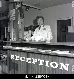 1975, historique, mécanicien de sexe masculin en manteau blanc avec logos de société, debout au comptoir de réception d'un garage, Croydon, Angleterre, Royaume-Uni. Banque D'Images