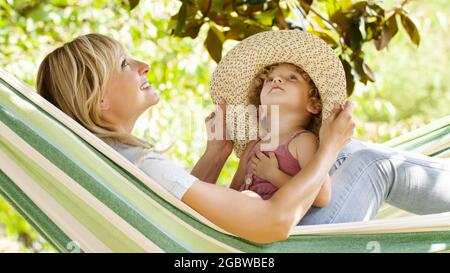 maman souriante et petite fille enfant yeux bleus avec des cheveux blonds bouclés, ensemble allongé sur le hamac dans le jardin vert de la maison, joue avec elle par Banque D'Images