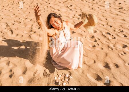 Belle femme caucasienne en robe blanche assise sur la plage jouant avec sable.Summer vacances concept. Banque D'Images