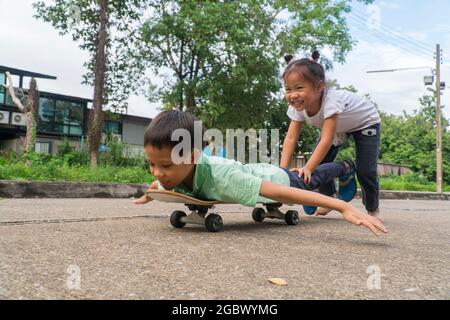 Petite fille adolescente açée jouant avec un frère poussant un garçon en avant sur le skateboard sur la voie publique. Enfants et divertissements. Enfants et sports con Banque D'Images