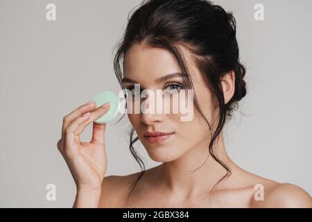 Jeune femme blanche avec les cheveux bruns tiré vers le haut appliquant le maquillage à l'aide de l'éponge de mélangeur de beauté isolée sur fond gris Banque D'Images