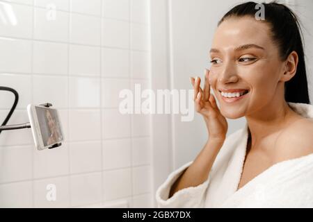 Jeune souriante jeune femme blanche beauté blogger dans bahtrobe faire une vidéo sur téléphone mobile dans la salle de bains appliquer le gommage du visage Banque D'Images