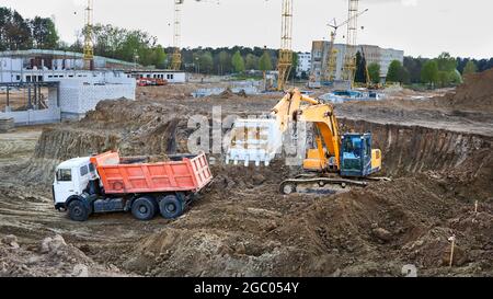 Excavatrice sur chenilles en cours de chargement du sable et du sol sur un camion minier de plusieurs tonnes sur un chantier de construction avec grues et ouvriers en arrière-plan Banque D'Images