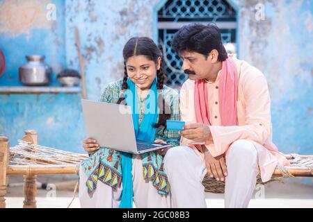 Père indien rural et jeune fille utilisant un ordinateur portable tout en étant assis sur un lit en bois traditionnel à l'extérieur de leur maison, famille heureuse regardant la scrore d'ordinateur Banque D'Images