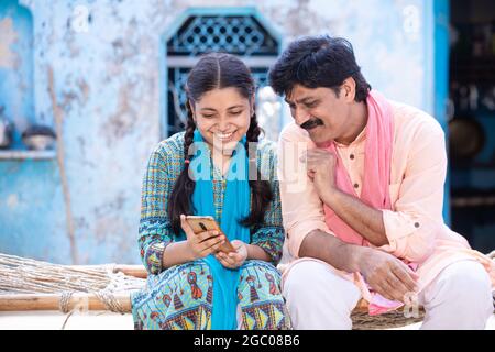 Père indien heureux et jeune fille utilisant un smartphone tout en étant assis sur un lit traditionnel, famille rurale heureuse regardant à l'écran mobile Banque D'Images