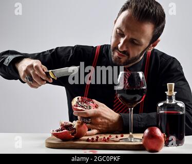 Homme barbu brutal en chemise noire avec cravate à rayures et bretelles assis à la table en mangeant du jus et du grenade frais Banque D'Images