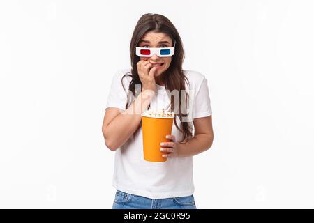 Concept de divertissement, de plaisir et de vacances. Portrait d'une jeune fille adorable et effrayée dans des lunettes 3d, mangeant du pop-corn, en butant et en piquant les doigts comme étant effrayés Banque D'Images