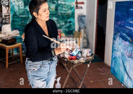 Peintre féminin concentré peint avec des couleurs bleues une pièce d'art abstraite dans son studio à la maison. Banque D'Images