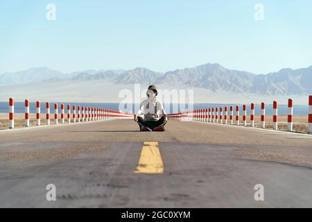 femme asiatique touriste assise au milieu d'une route vide avec lac et montagnes vallonnés en arrière-plan, jambe traversée. Banque D'Images