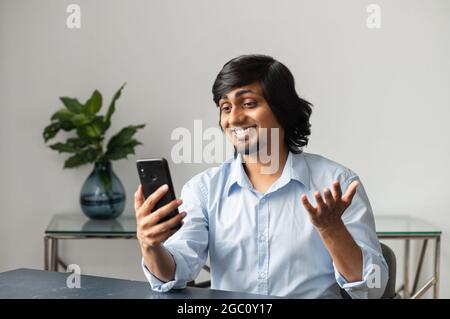 Homme indien souriant et amical effectuant des appels vidéo sur le smartphone, debout au bureau, tenant le téléphone à la main et parlant à distance, à l'aide de l'application mobile pour la communication virtuelle Banque D'Images