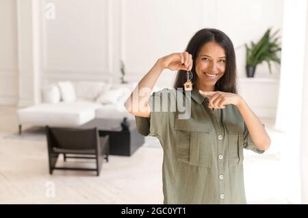 Concept de relocalisation. Photo de la femme indienne excitée qui se déplace dans une nouvelle maison ou un nouvel appartement. Joyeuse femme de course mixte regardant l'appareil photo et pointe le doigt sur les clés avec le porte-clés dans sa main Banque D'Images