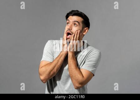 Choqué jeune homme caucasien avec les mains sur les joues qui gazouchent et regardant vers le haut dans le fond gris clair isolé studio Banque D'Images