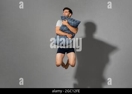 Portrait d'un jeune homme énergique et frais portant des vêtements de nuit tenant un oreiller et sautant en plein air après un bon sommeil le matin, studio Banque D'Images