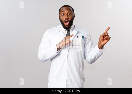 J'ai impressionné le médecin afro-américain masculin de bonne qualité en manteau blanc, pointant vers le coin supérieur droit, éveiller et affamer la caméra excitée et émerveillée, dire Banque D'Images