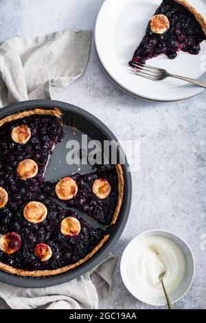 Tarte aux bleuets en tranches dans son moule avec une tranche sur une assiette avec une fourchette et un petit bol de crème fouettée. Banque D'Images