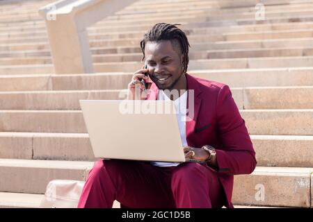 souriant homme noir travaille avec ordinateur portable et téléphone assis sur les escaliers de la ville, la technologie et le concept de travail à distance Banque D'Images