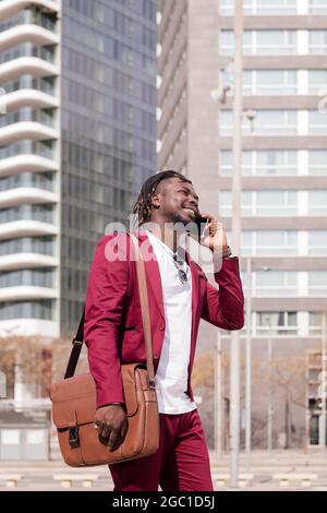 photo verticale d'un homme d'affaires africain souriant avec porte-documents marche à travers la ville financière centre-ville parlant au téléphone, concept de la technologie et c Banque D'Images